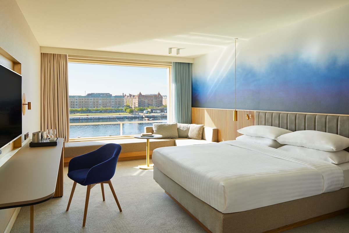 Habitación de hotel con una cama grande, un escritorio con televisor, una silla azul y vista a un paisaje urbano por la ventana.