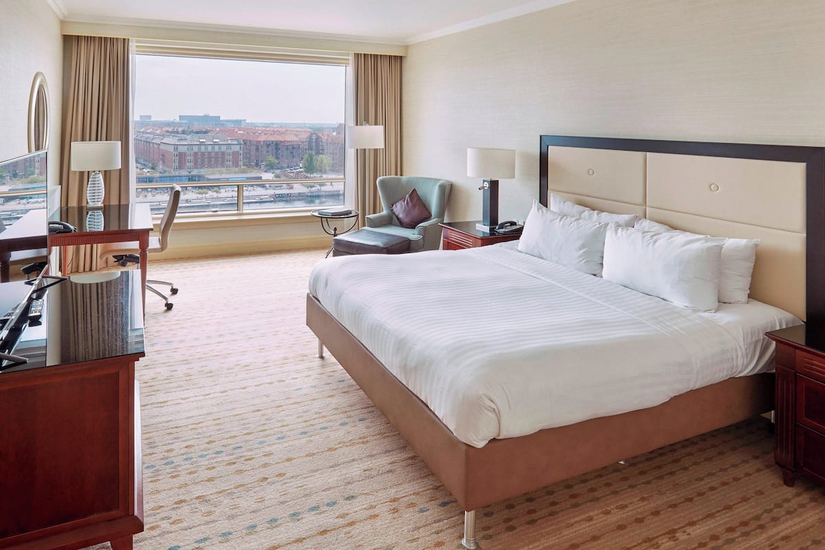 Habitación de hotel con una cama grande, un escritorio con teléfono, una silla y vistas a la ciudad a través de un amplio ventanal.
