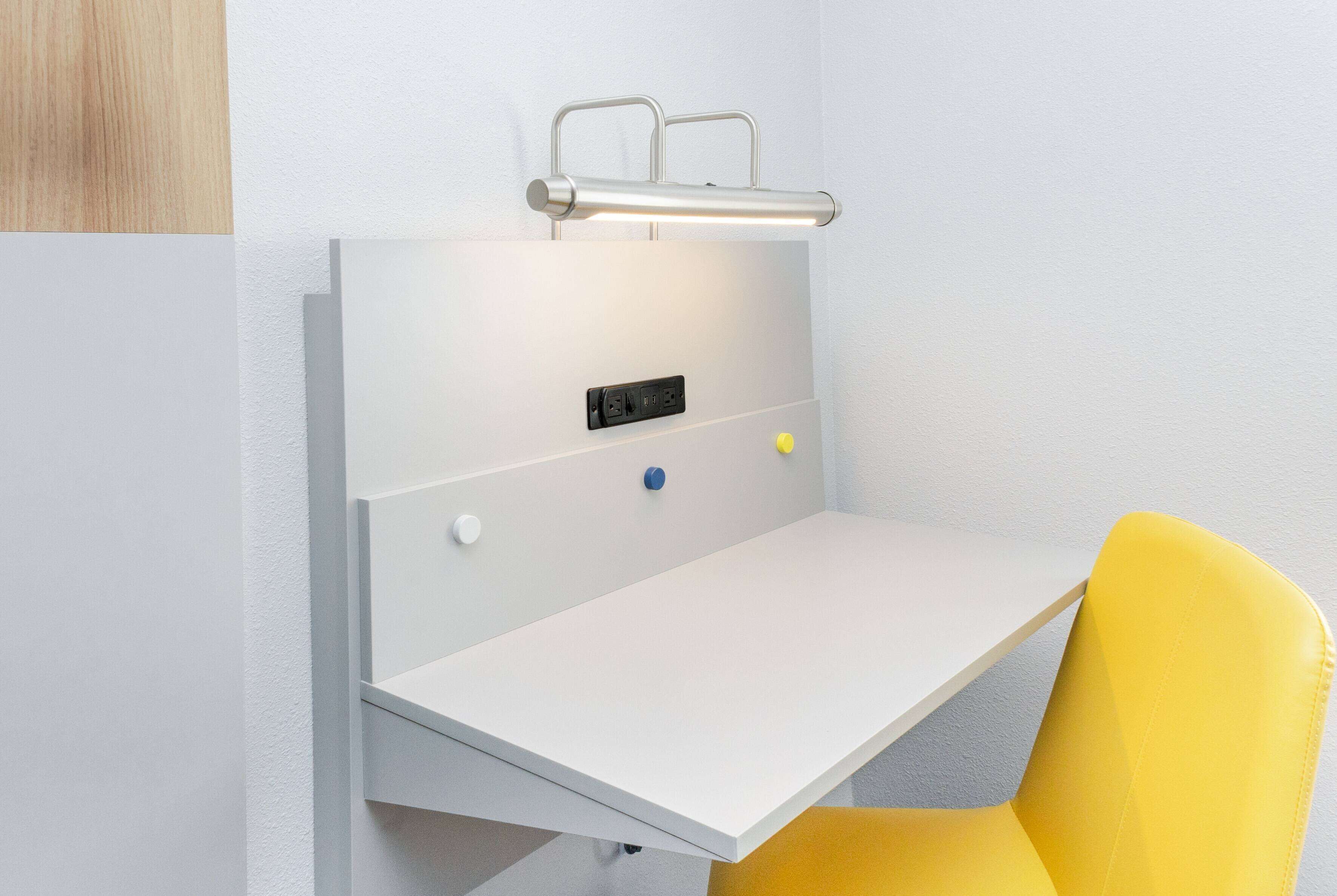 A white desk with a yellow chair, a light fixture above, and a power outlet with a blue switch.