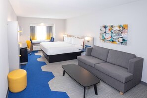 A hotel room with a bed, a grey sofa, a small table, a yellow ottoman, and a painting on the wall.