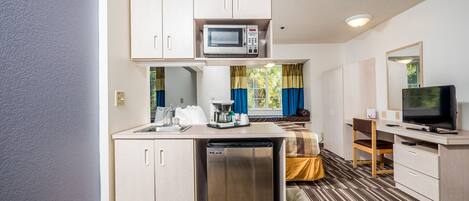 A compact hotel room with a kitchenette, a bed, a television, and a window with curtains.