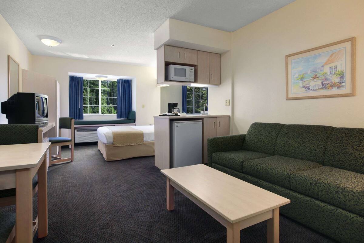 A compact hotel room with a bed, a green sofa, a small kitchenette, a television, and a window with curtains.