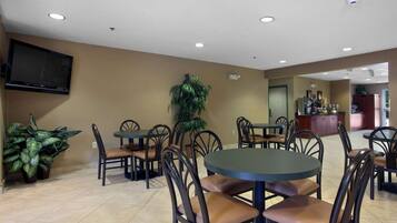 A dining area with round tables and chairs, a flat-screen TV mounted on the wall, and potted plants.