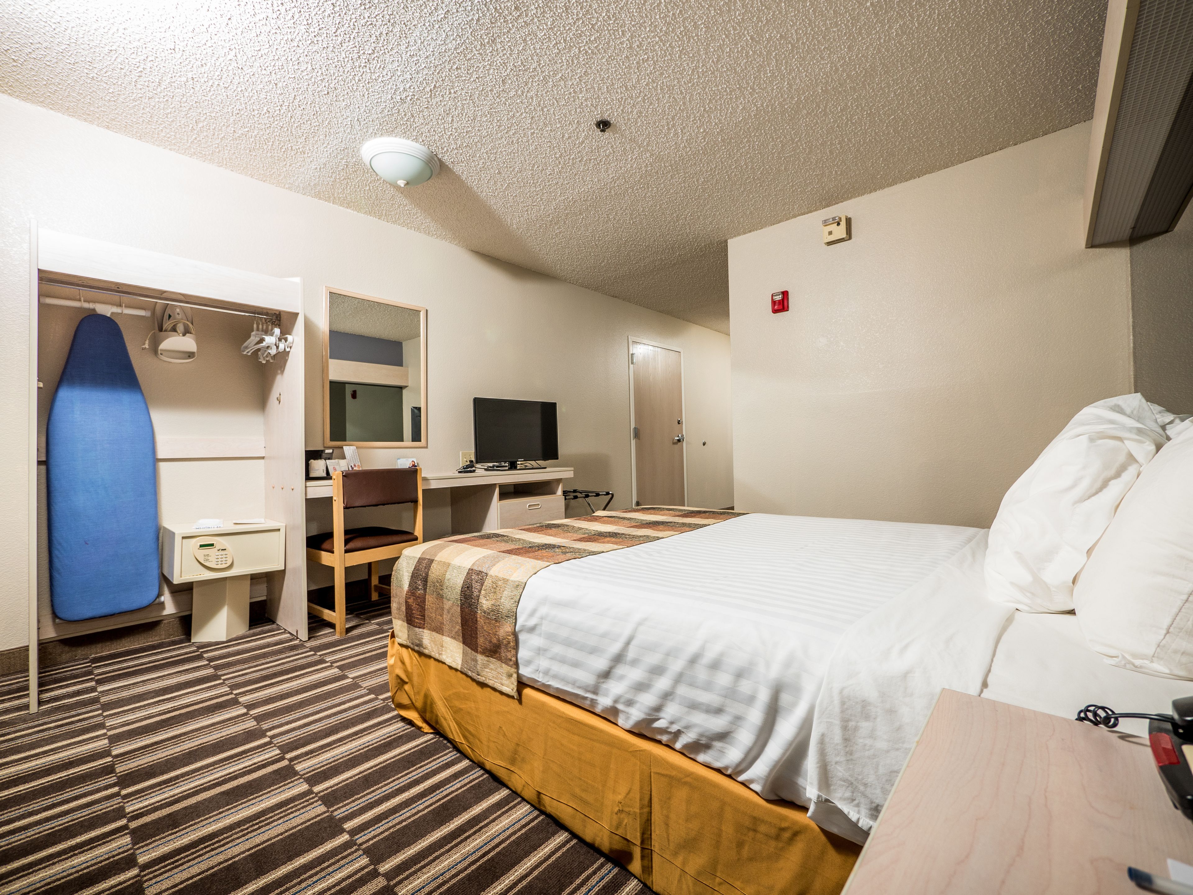 Une chambre d'hôtel avec un lit, un bureau avec un ordinateur, une chaise, un miroir, une planche à repasser bleue et un sol recouvert de moquette.