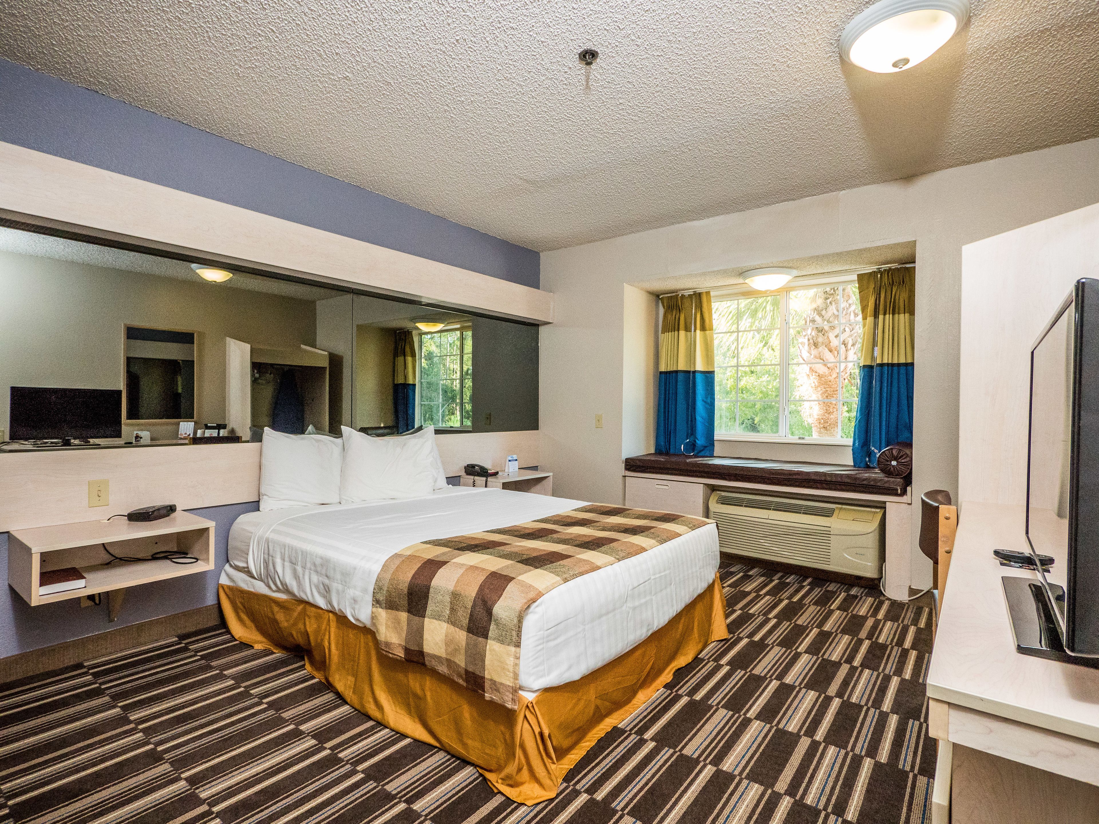 A hotel room with a bed, a desk, a mirror, a window with curtains, and a television.