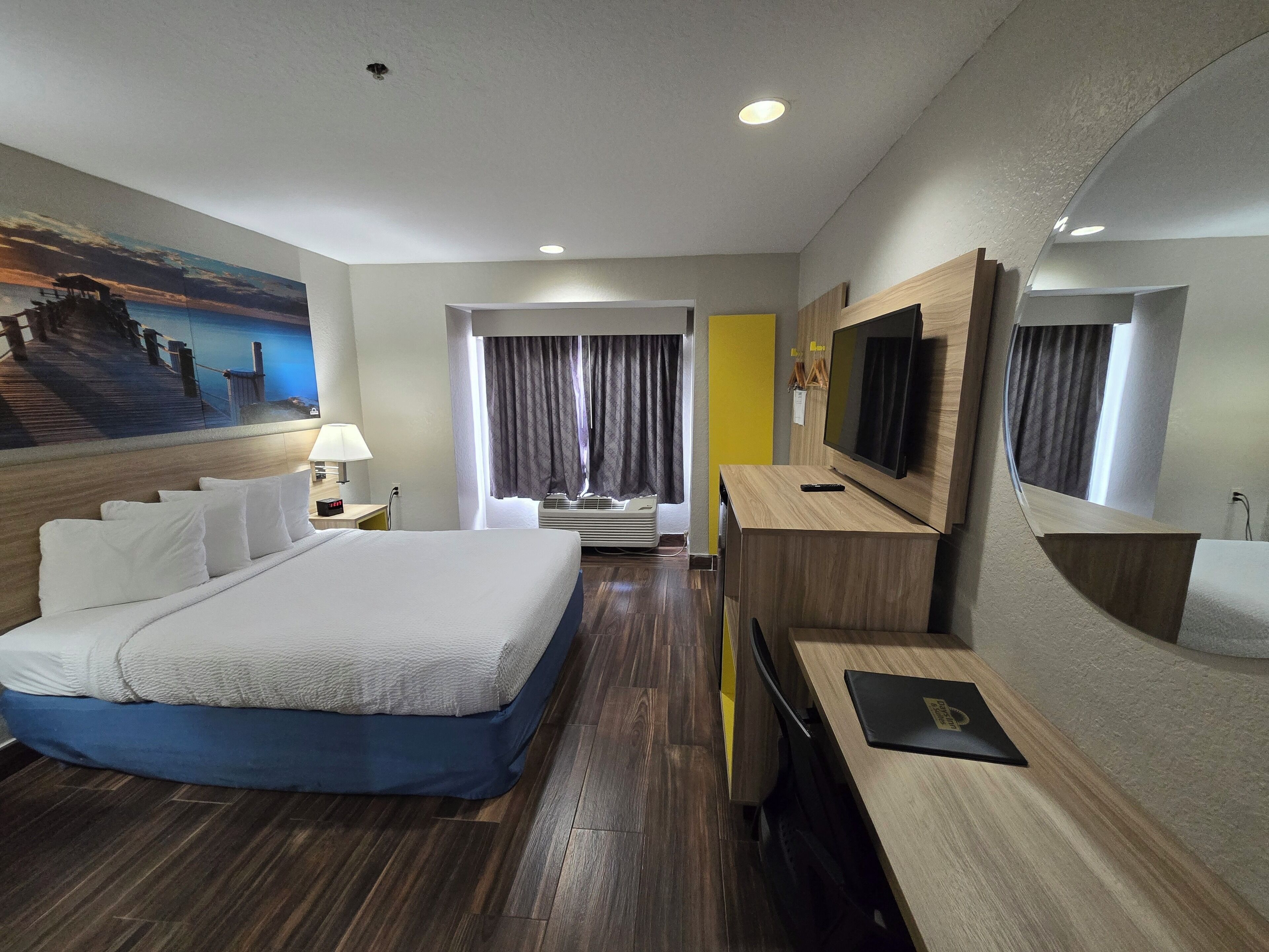 Quarto de hotel com uma cama grande, uma escrivaninha com televisão, um quadro na parede e uma janela com cortinas.