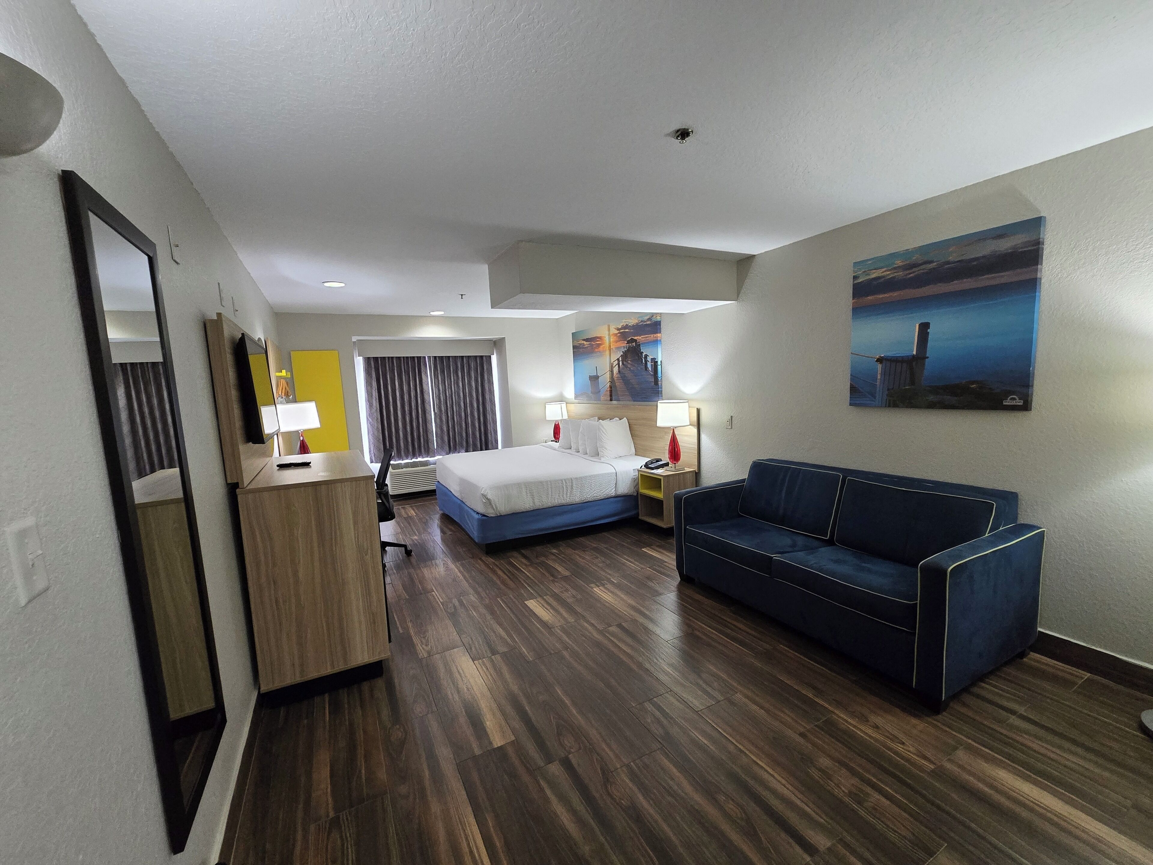 A hotel room with a bed, a desk, a chair, a blue sofa, and a painting on the wall.