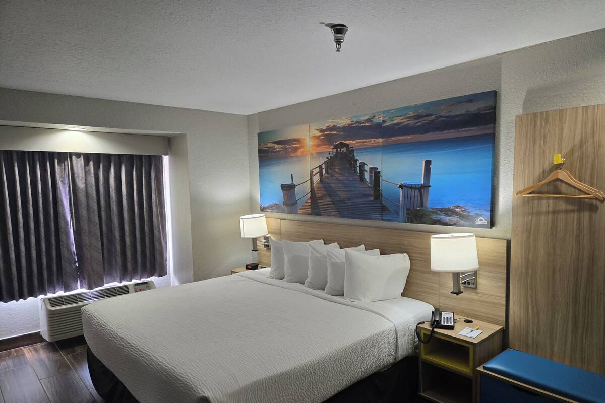 Habitación de hotel con una cama grande, dos lámparas de noche, un cuadro de una puesta de sol sobre el agua, cabecera de madera, perchero y un escritorio con teléfono.