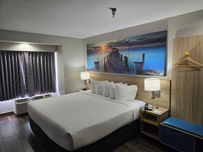 Une chambre d’hôtel avec un grand lit, deux lampes de chevet, un tableau représentant un coucher de soleil sur l’eau, une tête de lit en bois, un porte-manteau suspendu et un bureau avec un téléphone.