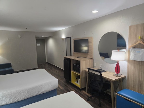 Quarto de hotel com duas camas, uma escrivaninha com computador, televisão, espelho e uma pequena cozinha.