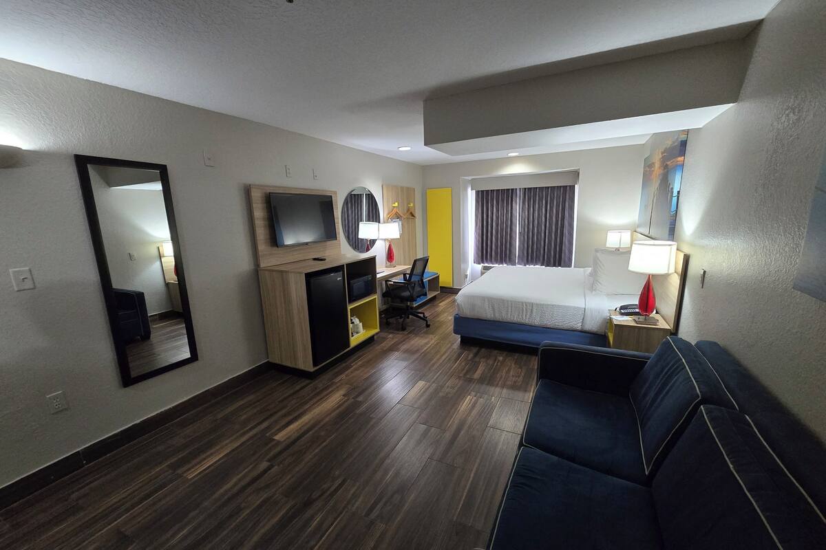 Habitación de hotel con cama, escritorio, silla, televisor, espejo y ventana con cortinas.