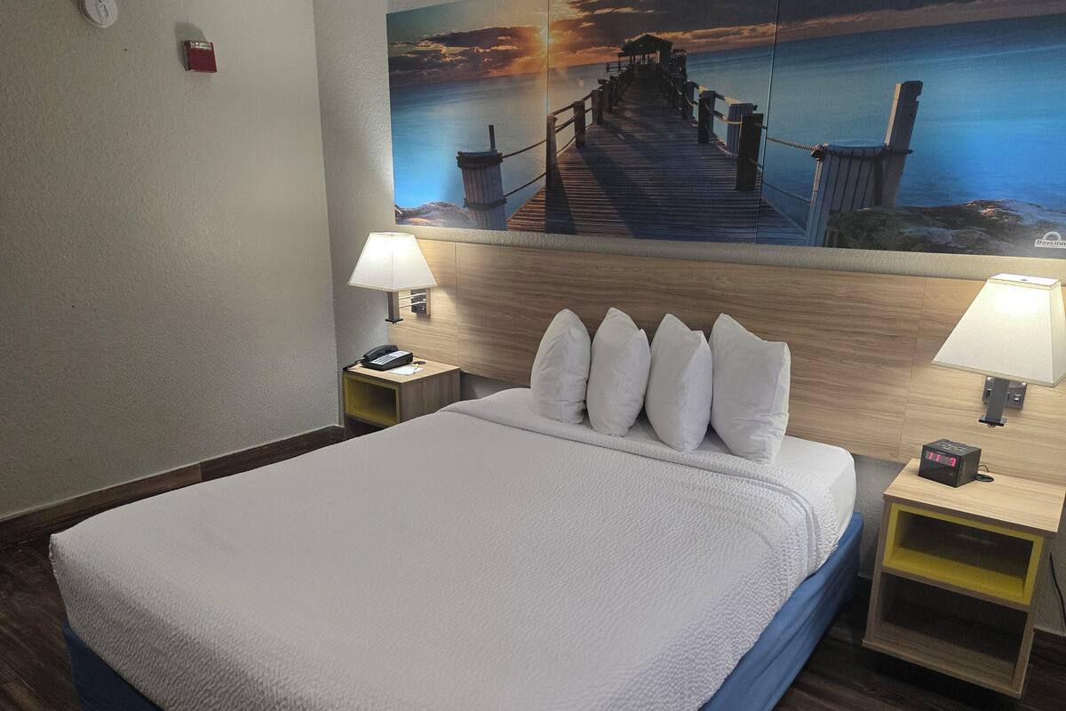 Habitación de hotel con una cama grande, dos mesitas de noche, una lámpara de pared, un teléfono y un cuadro de un muelle.