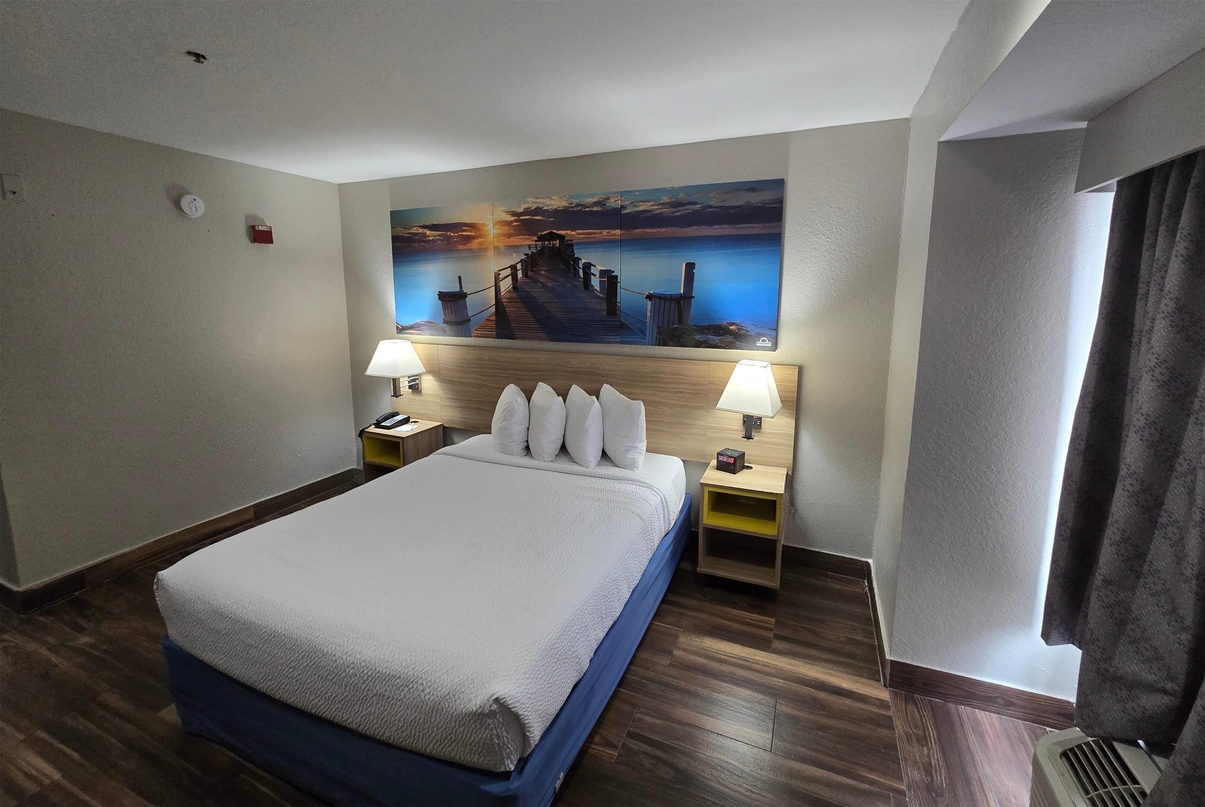 Une chambre d’hôtel avec un lit, deux tables de chevet, une petite table, un tableau représentant un coucher de soleil sur l’eau et une fenêtre avec des rideaux.