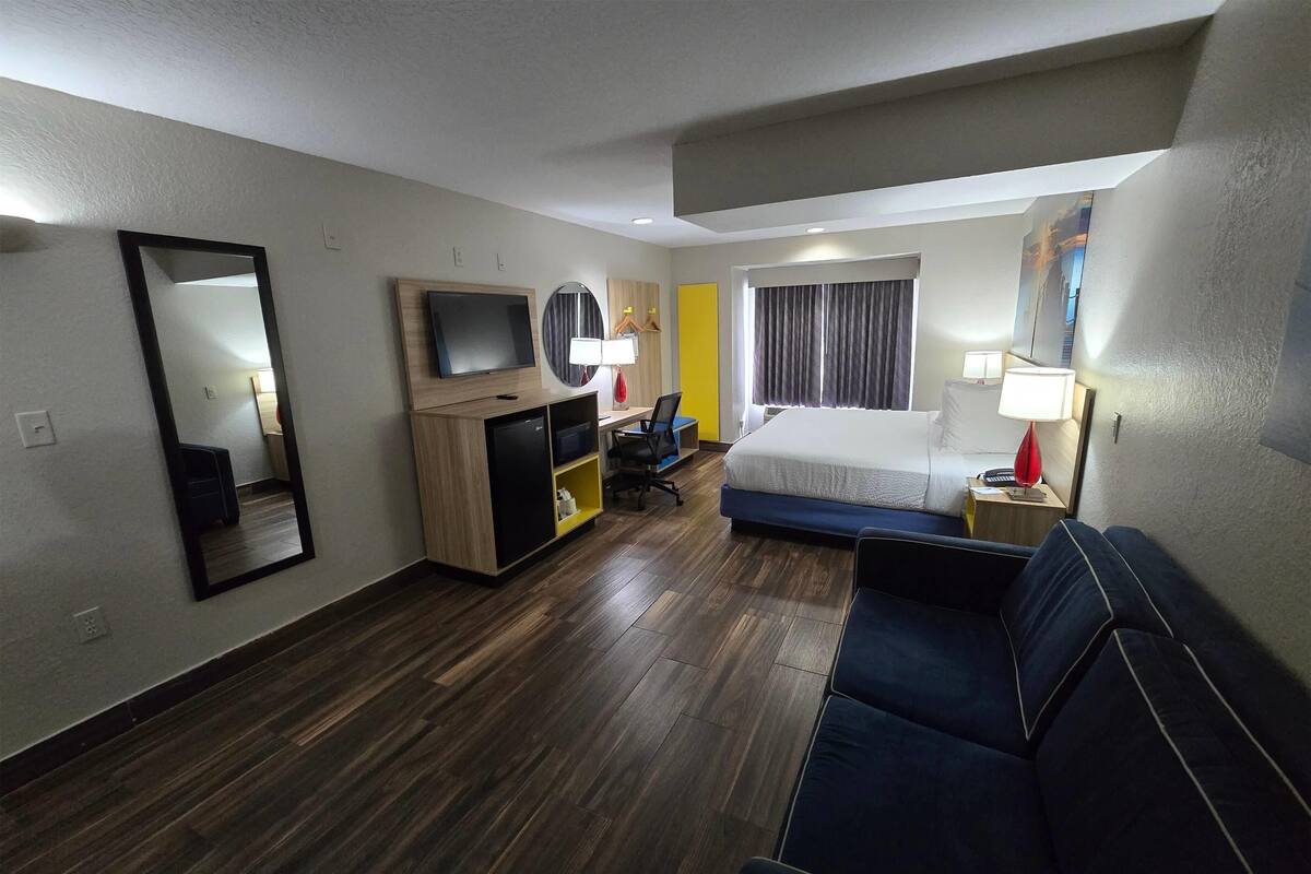 Habitación de hotel con cama, sofá, escritorio con silla, televisor y espejo.