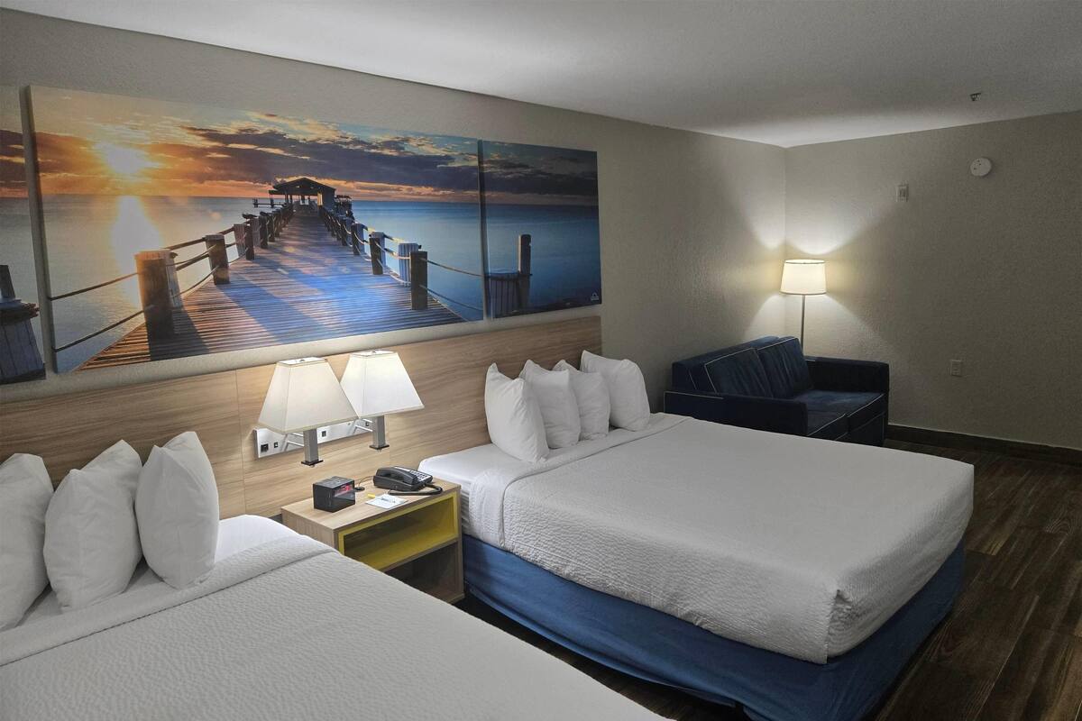 Habitación de hotel con dos camas, un escritorio con lámpara, una silla y un cuadro grande de un muelle al atardecer.