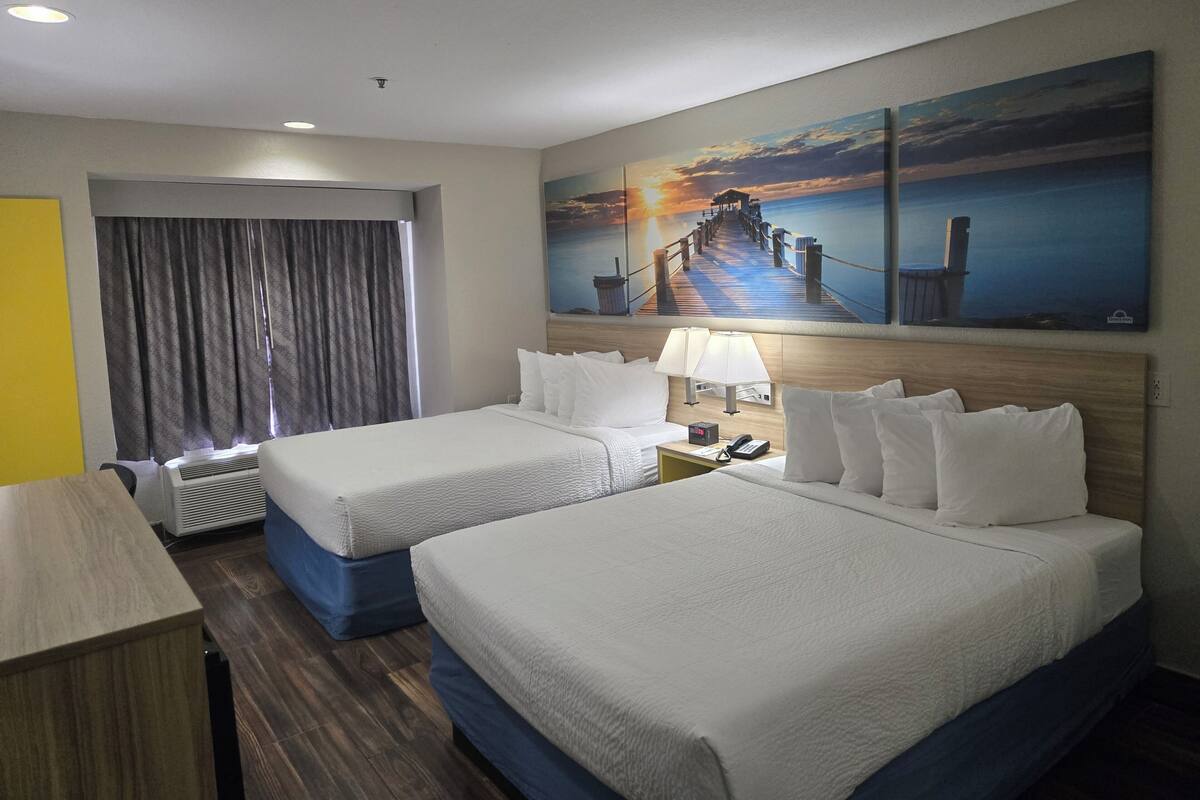 Habitación de hotel con dos camas, un escritorio, una lámpara y un cuadro de un atardecer sobre un muelle.