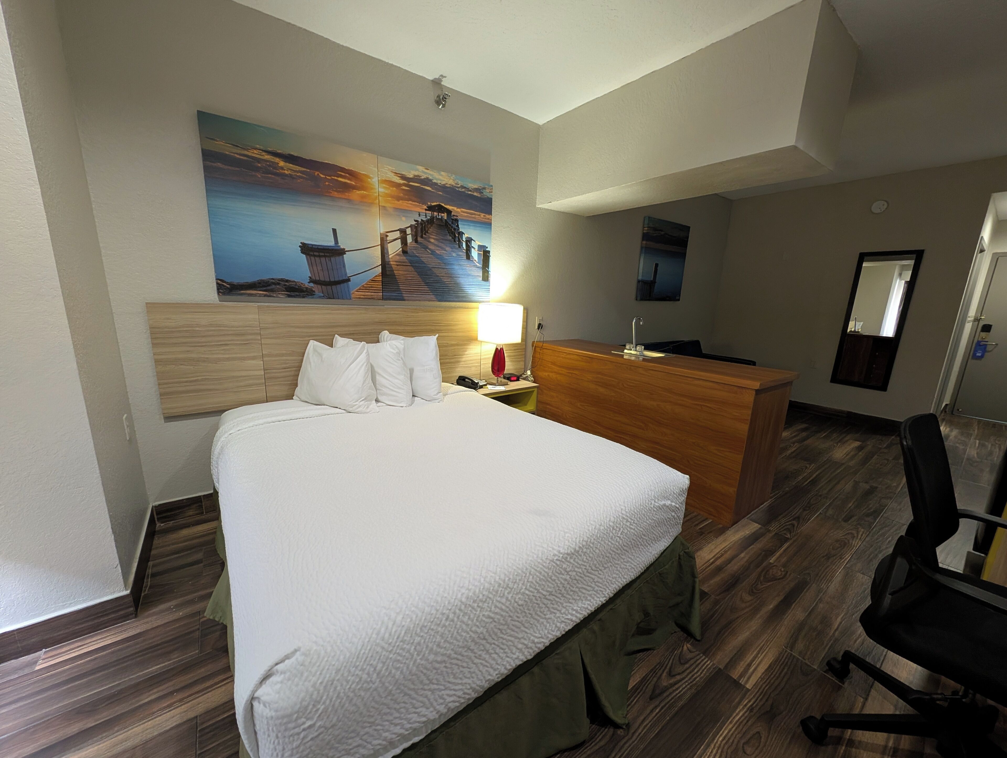 Ein Hotelzimmer mit Bett, Schreibtisch, Stuhl, einem Gemälde mit Sonnenuntergang über Wasser und Holzboden.