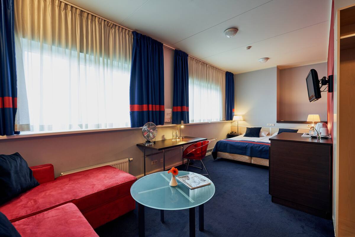 A hotel room with a red sofa, a glass table with a vase, a desk with a chair, a bed, and a TV mounted on the wall.