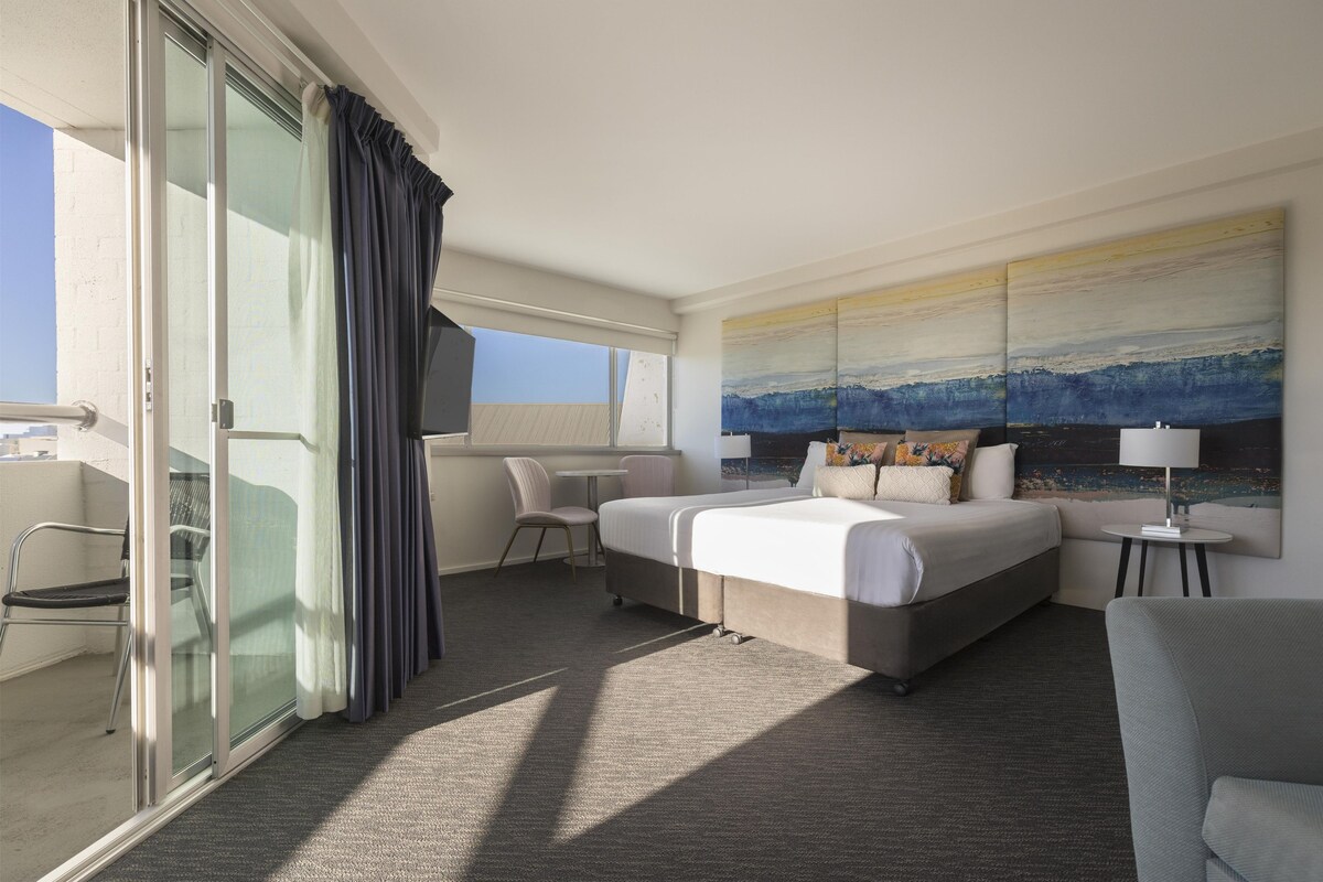 A hotel room with a bed, a chair, a small table, and a large window with curtains.