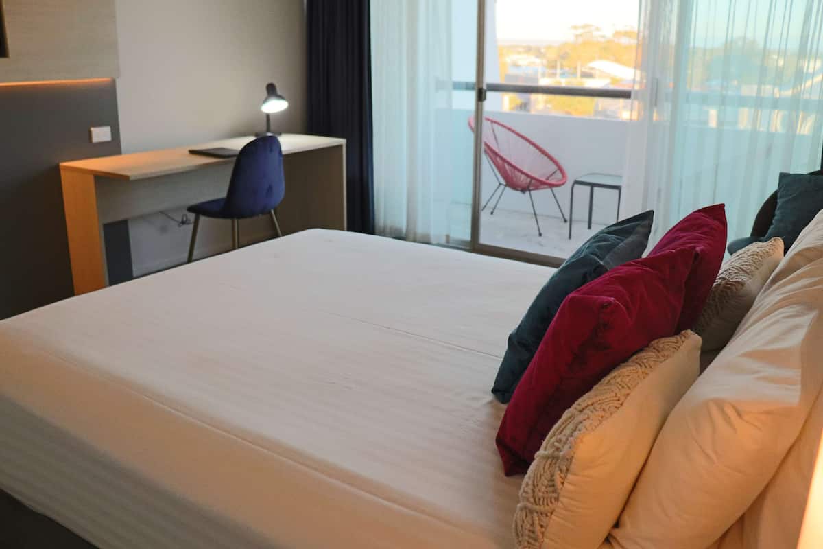 A hotel room with a bed, a desk, and a chair. There is a view of the outside through large windows.