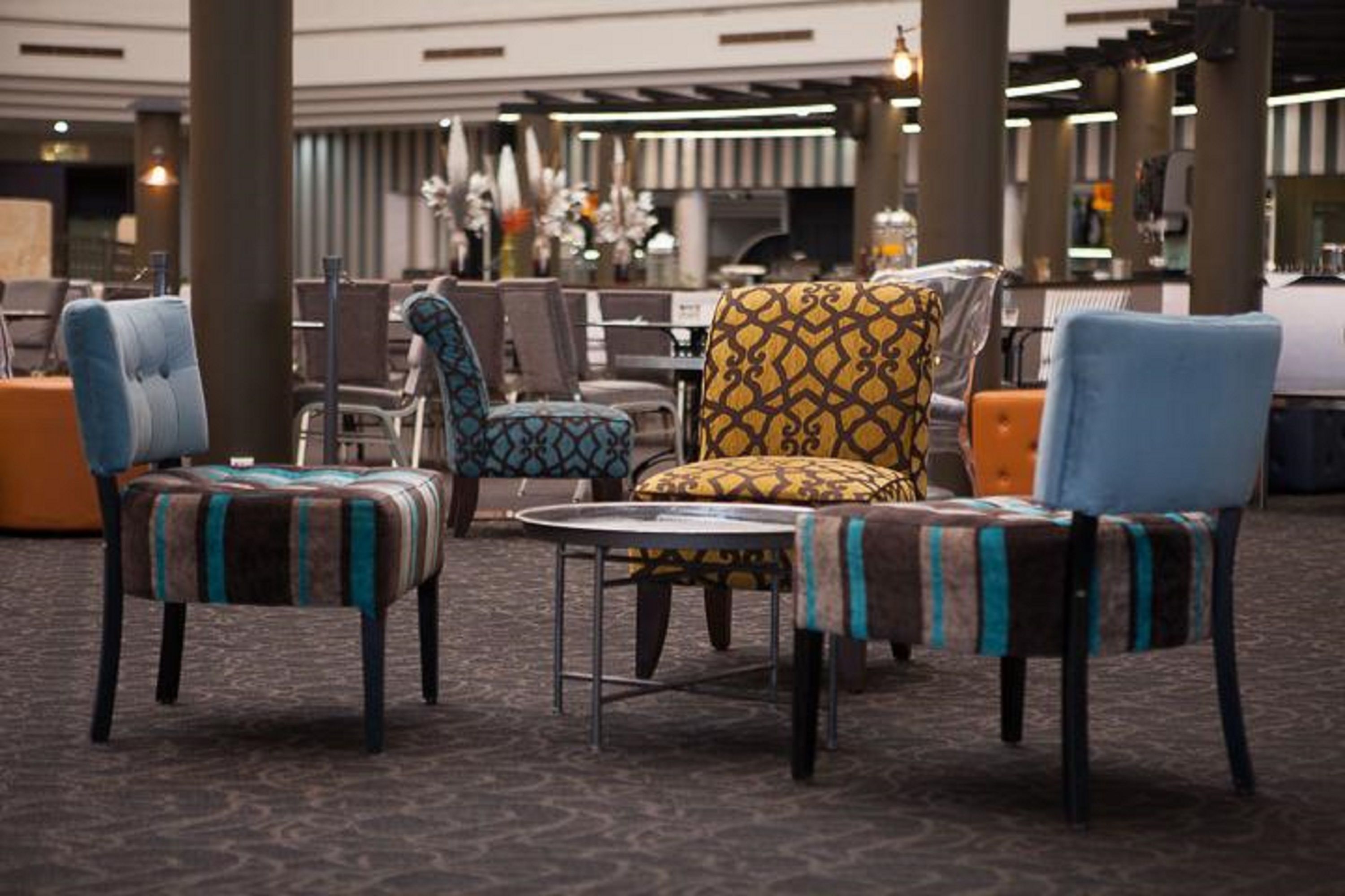 A hotel lobby with a seating area featuring patterned armchairs and ottomans, a round table, and a decorative wall with hanging lights.