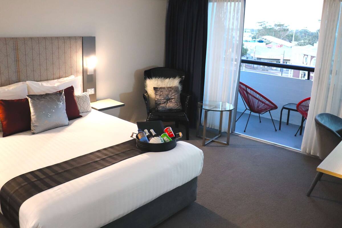 A hotel room with a large bed, a chair, a small table, and a balcony with red chairs.