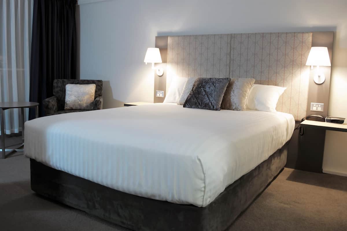 A large bed with a headboard, two bedside lamps, a small table, and a chair.