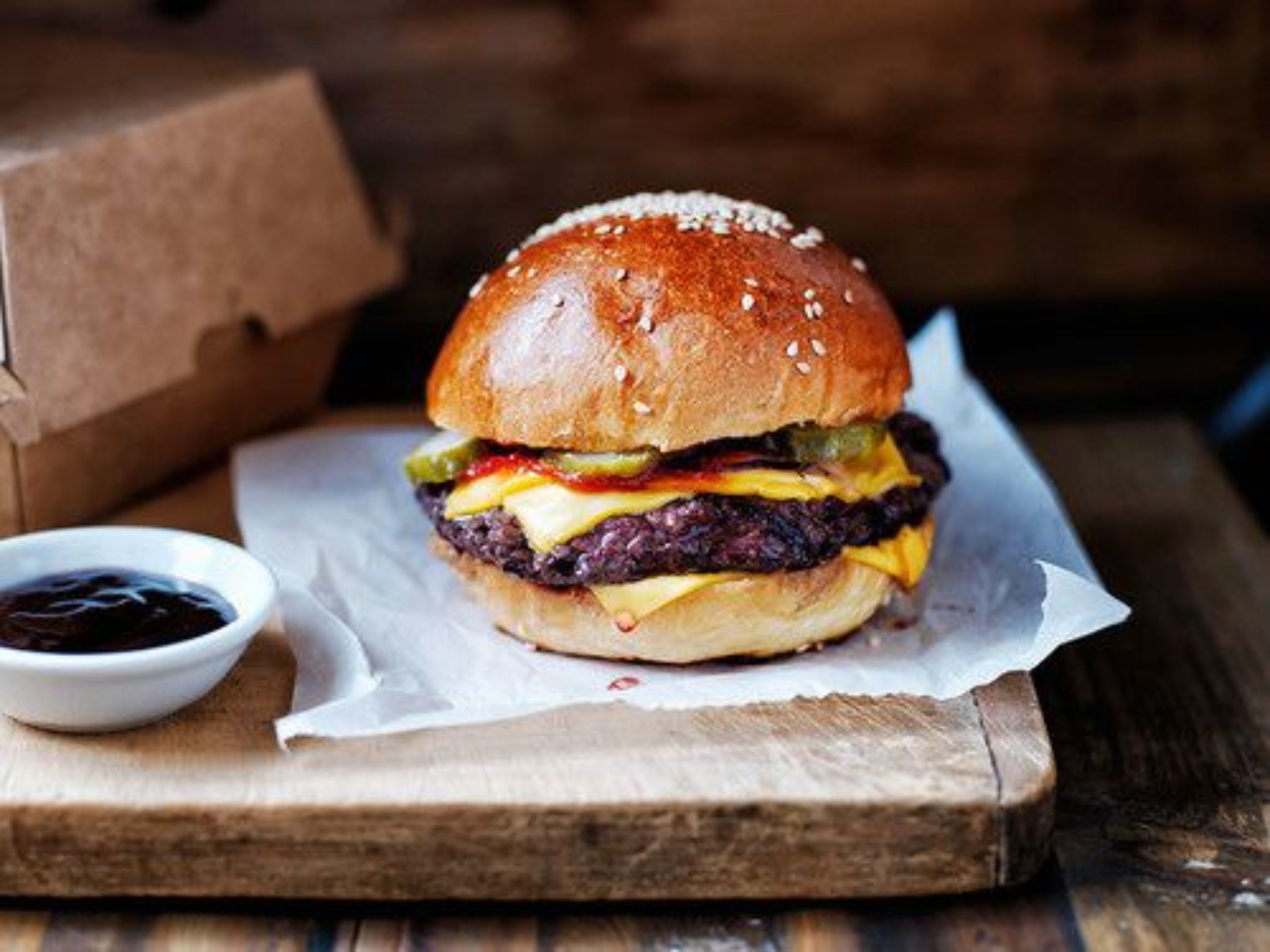 Hamburguesa con pan de sésamo, queso, pepinillos y una hamburguesa de carne, servida sobre una tabla de madera con un pequeño recipiente de salsa.
