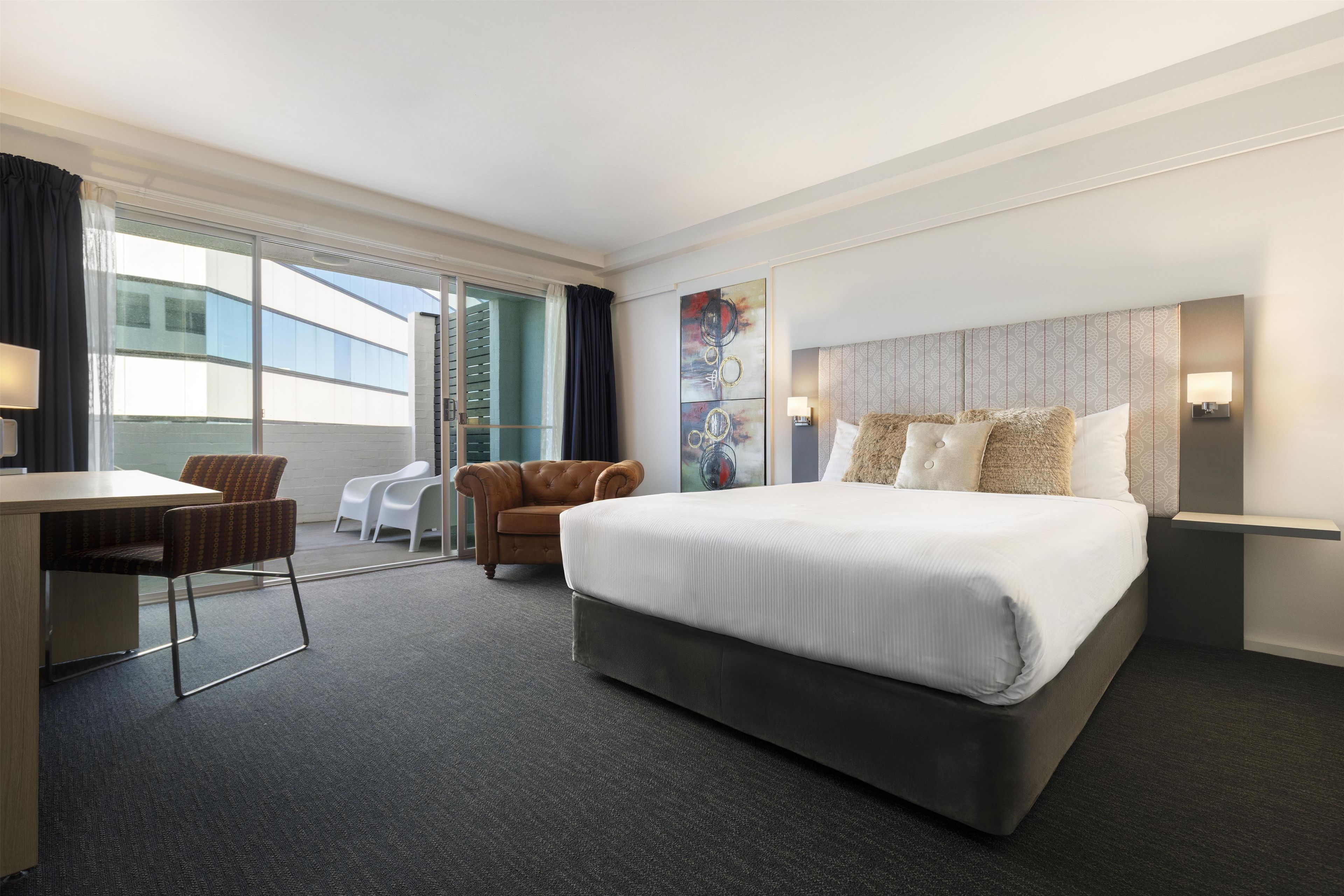 A hotel room with a large bed, a desk, two chairs, a view of the outside, and a modern headboard.