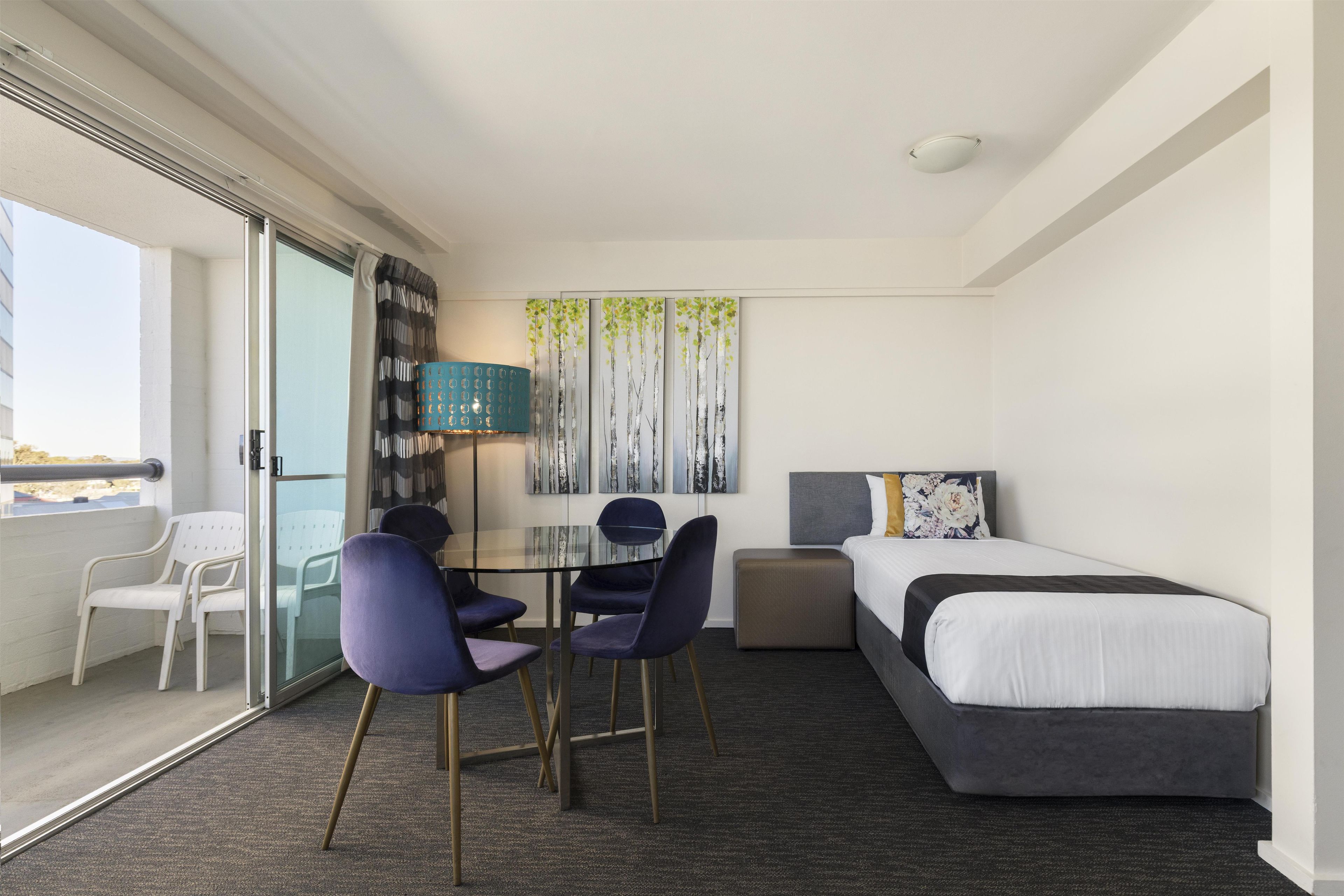 Een hotelkamer met een bed, twee stoelen, een klein tafeltje, een lamp en een schilderij aan de muur.