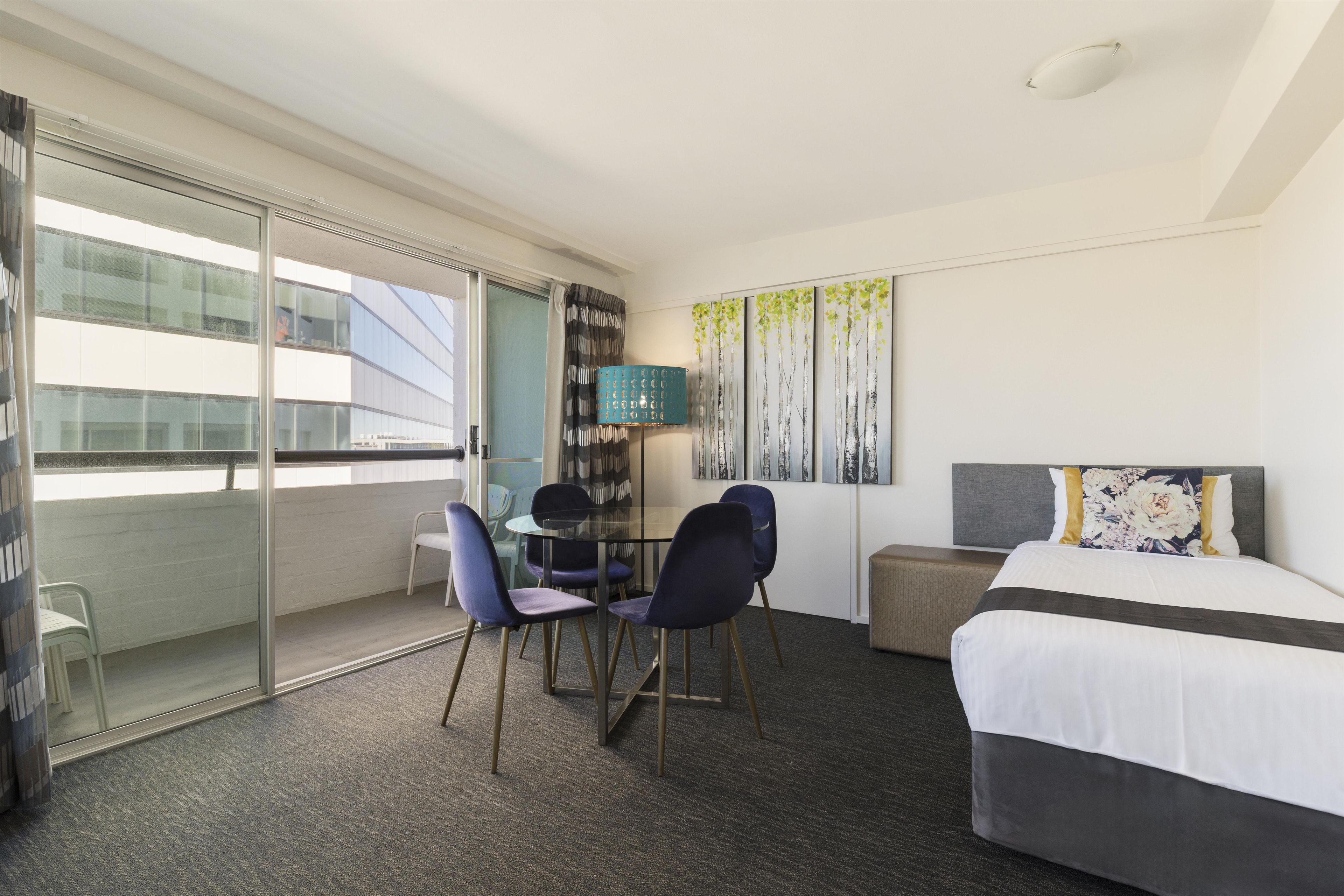 A hotel room with a bed, a desk with chairs, a small table, a sofa, and a large window.
