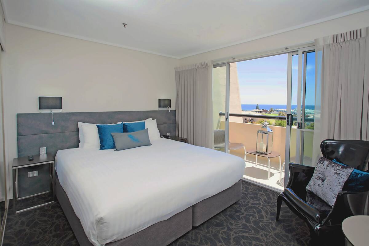 A hotel room with a large bed, a chair, a small table, and a balcony with a view of the ocean.