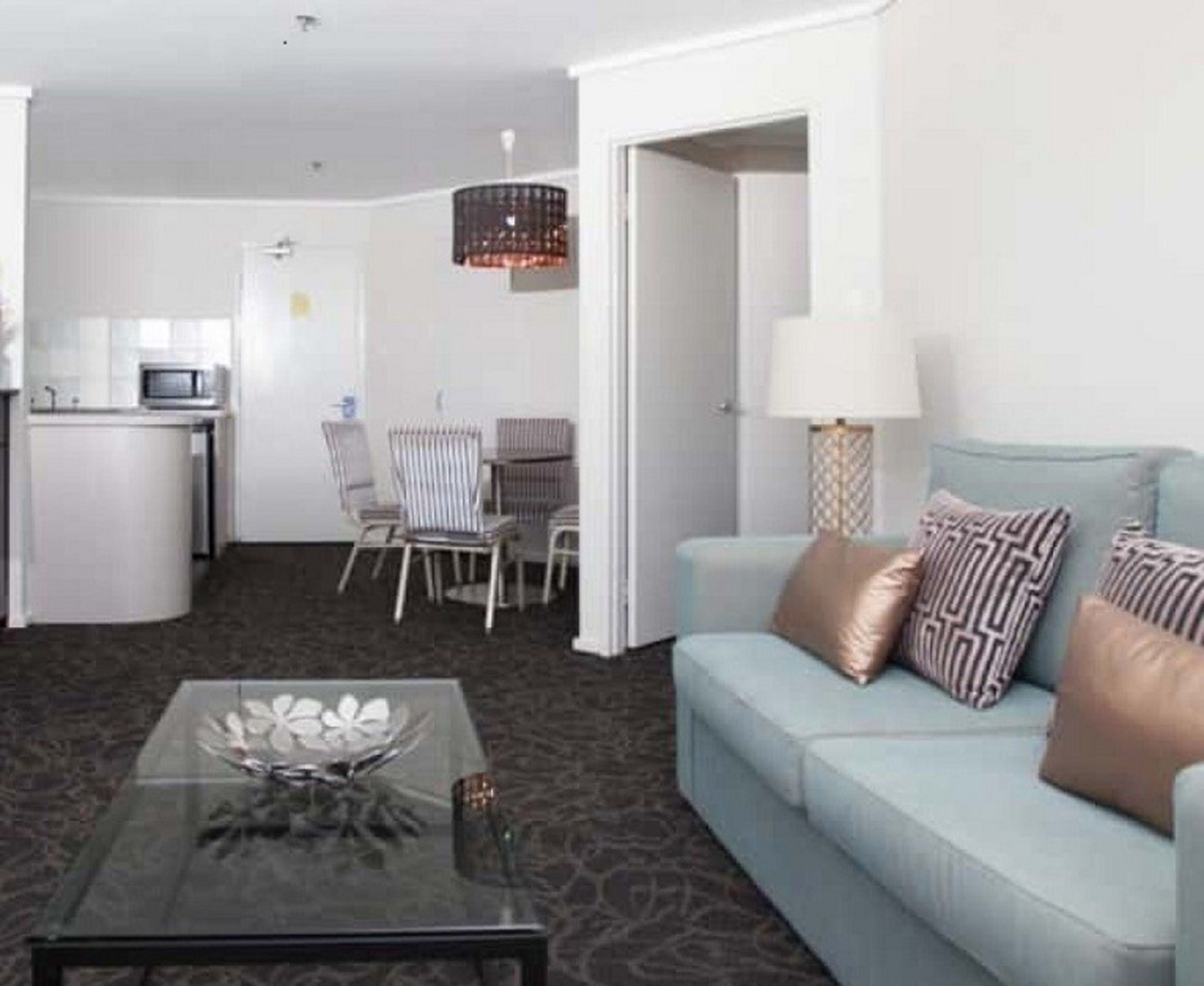 A compact living space with a sofa, a glass coffee table, a dining area with chairs, a kitchen area with a microwave, and a hanging light fixture.