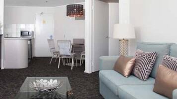 A compact living space with a sofa, a glass coffee table, a dining area with chairs, a kitchen area with a microwave, and a hanging light fixture.