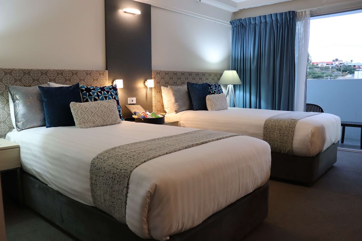 A hotel room with two beds, a nightstand, a lamp, and a view of the outside.