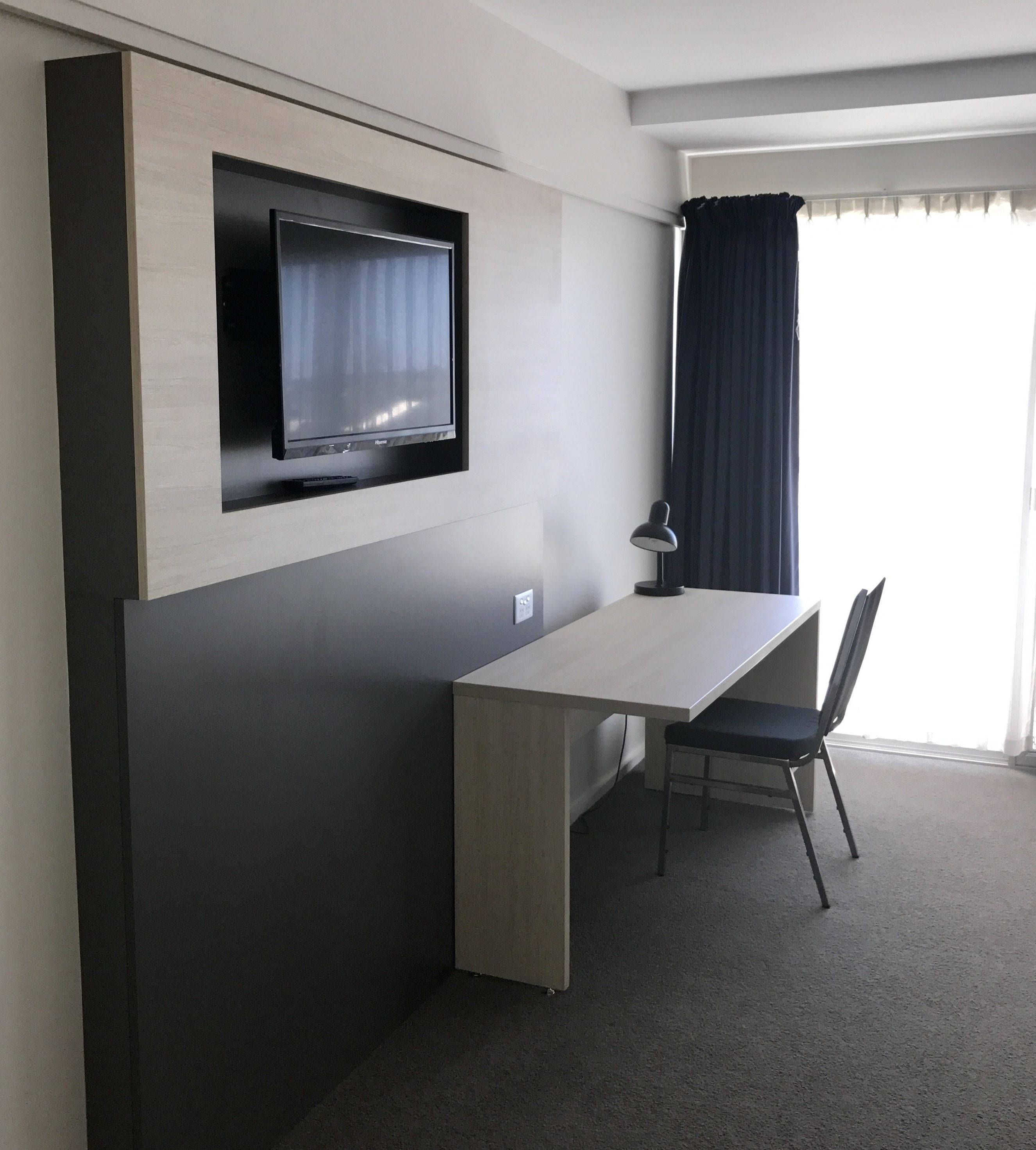 A modern hotel room with a built-in TV, a desk, a chair, and a lamp.