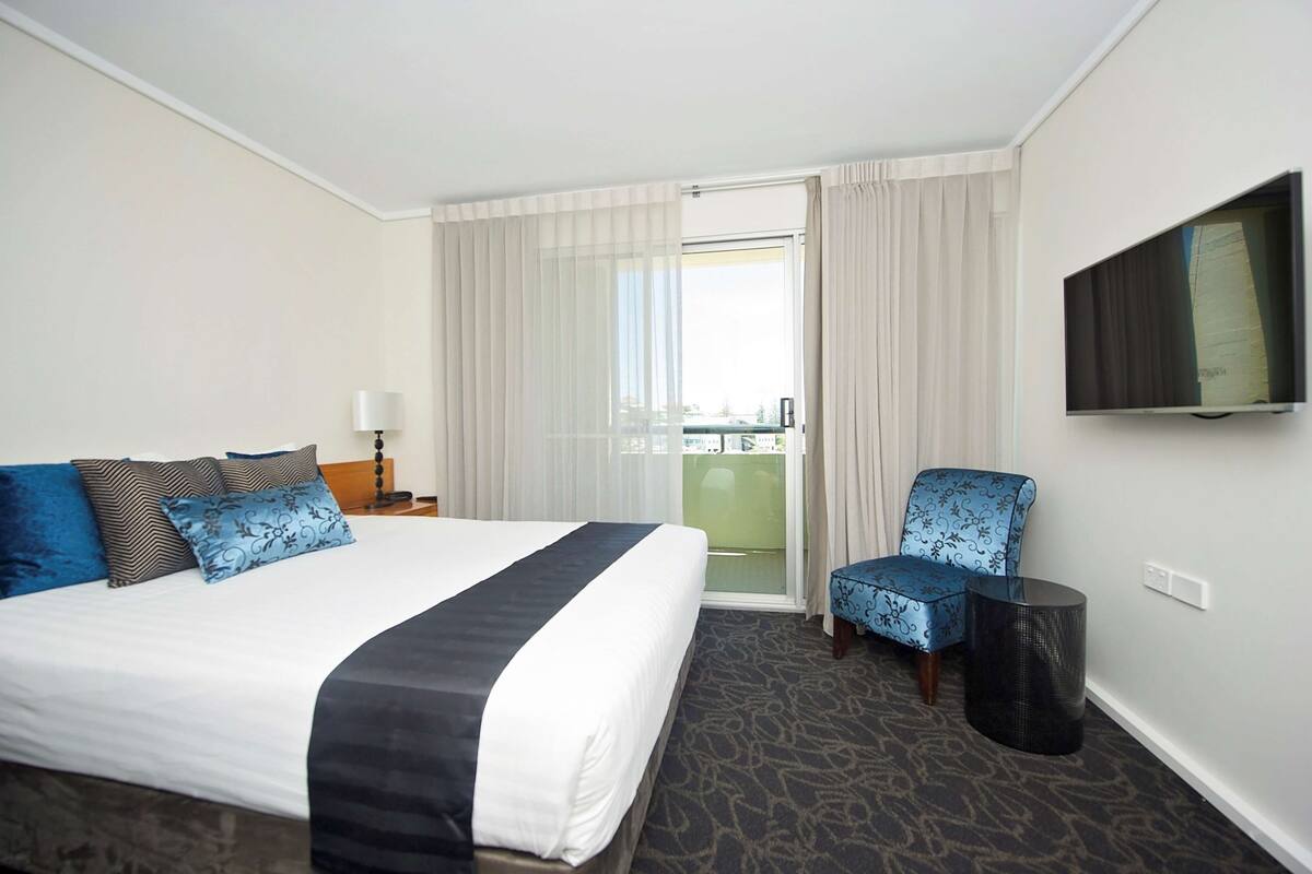 A hotel room with a large bed, a chair, a TV, and a balcony with curtains.