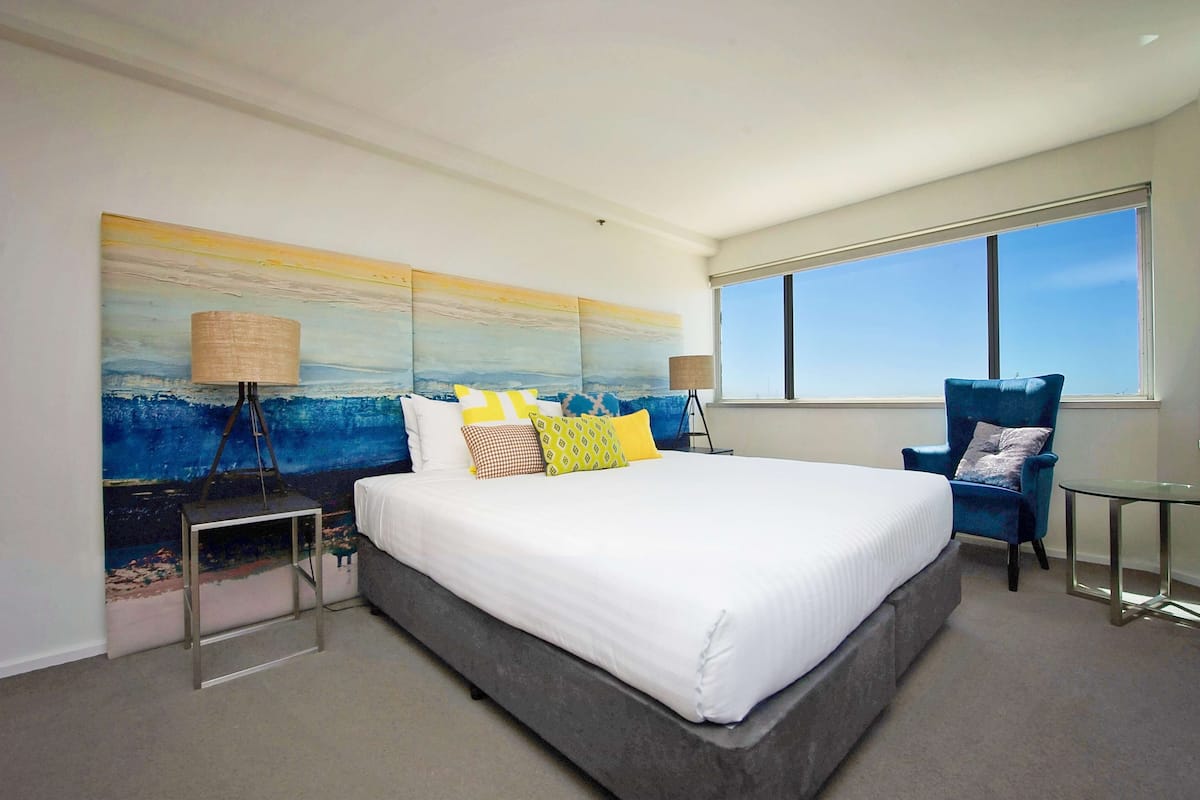 A modern hotel room with a large bed, a blue armchair, a small table, and a large window offering a view of the sky.