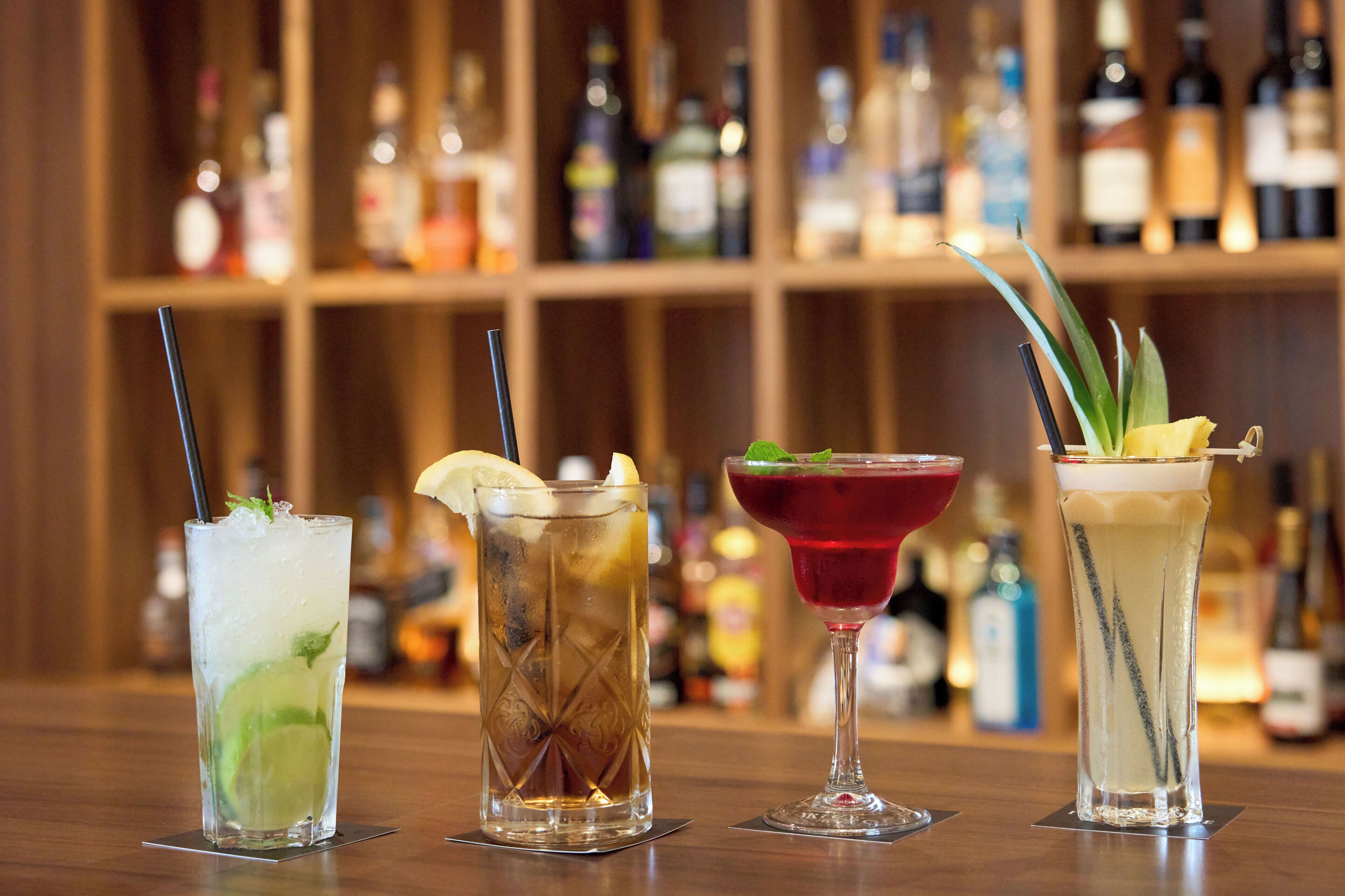 Quatre cocktails différents sont présentés sur le comptoir d’un bar, avec en arrière-plan des étagères remplies de bouteilles de toutes sortes.