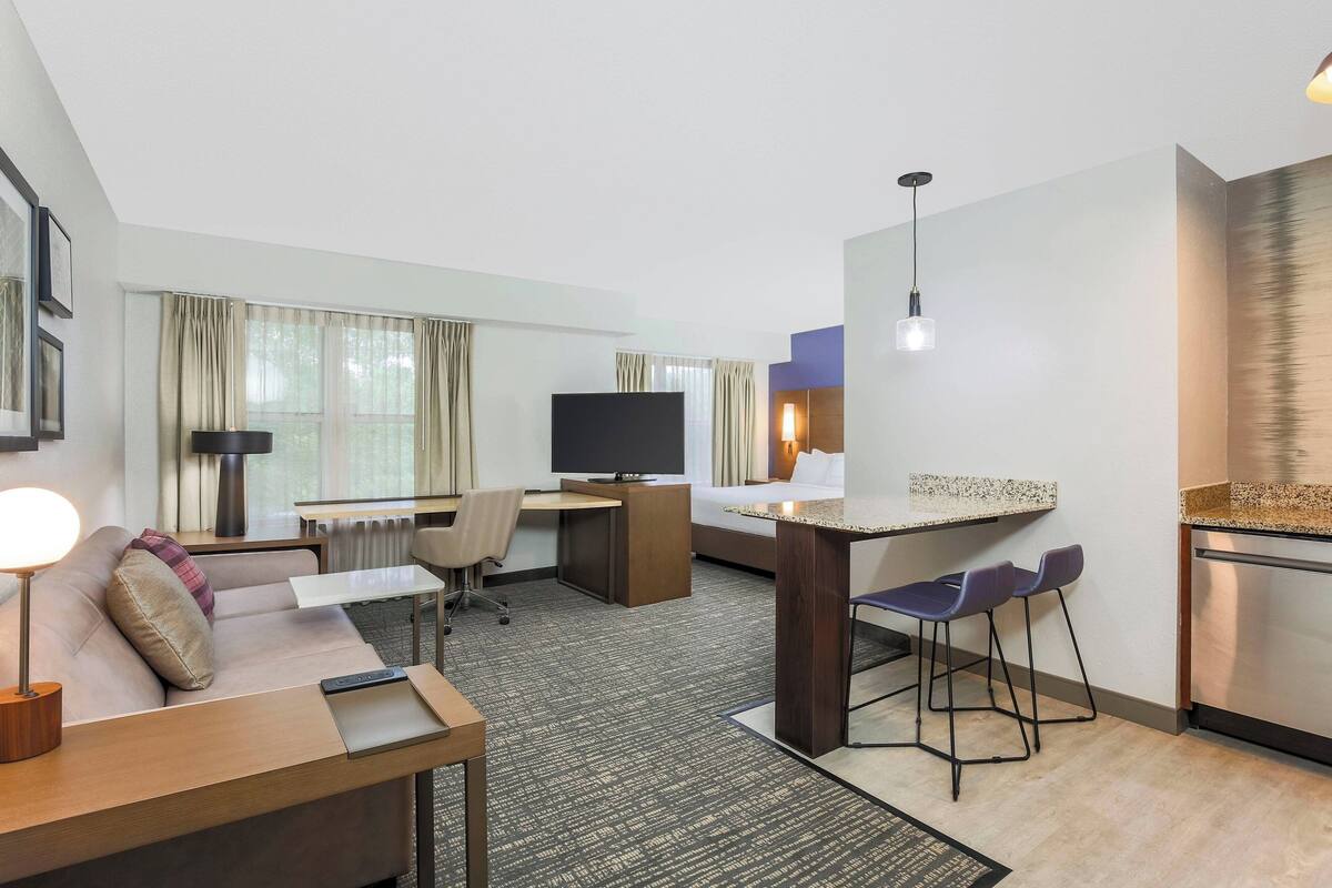 Moderni hotellihuone, jossa on sohva, työpöytä, televisio ja pieni keittiö.