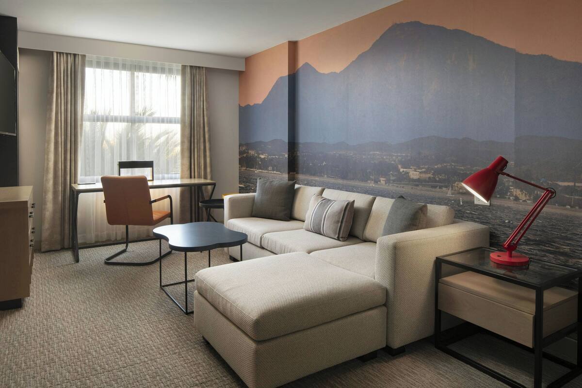 A hotel room with a sofa, a small table, a desk, and a large window with curtains.