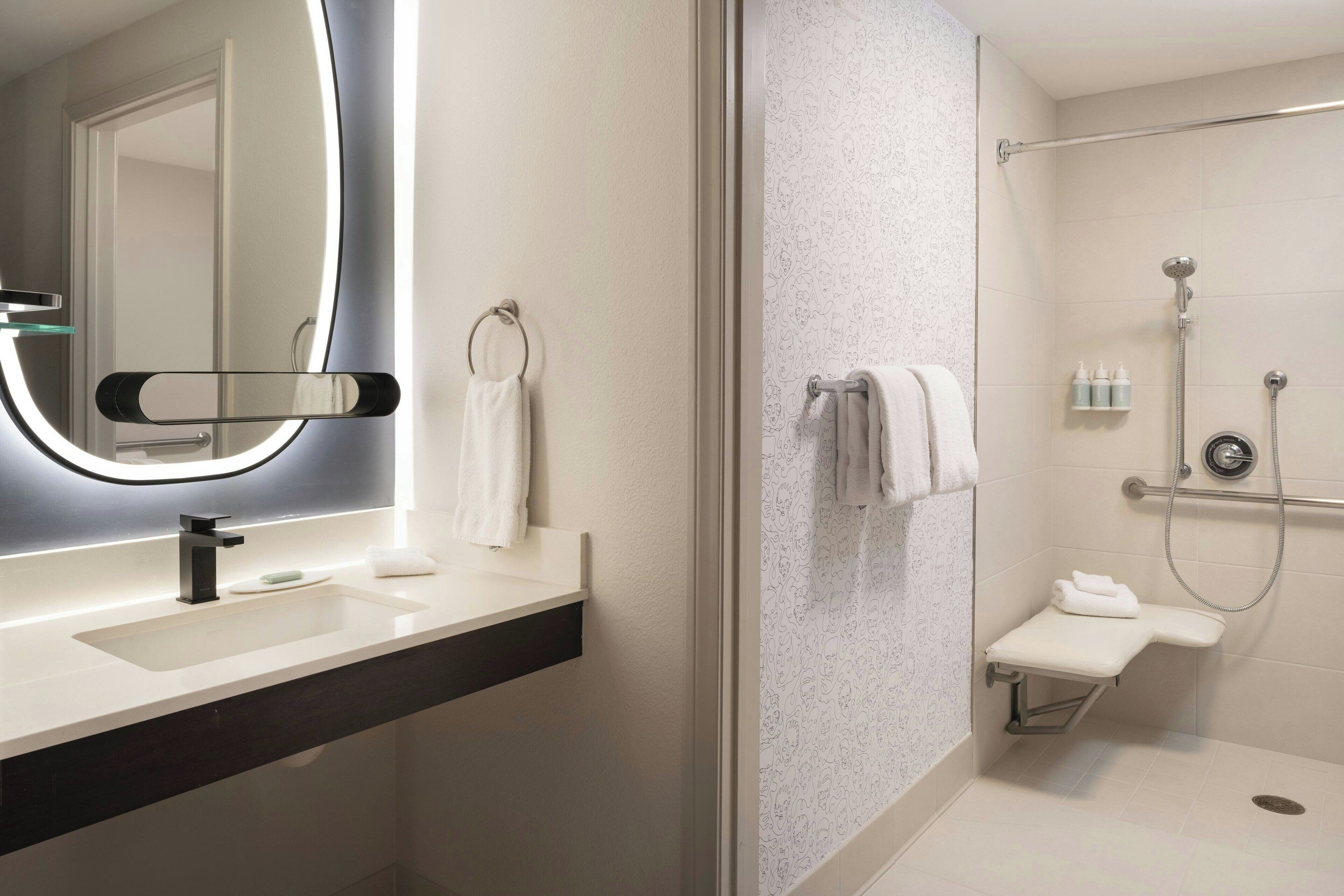 Une salle de bain moderne avec un grand miroir, un lavabo, une douche équipée d’un pommeau de douche à main et un porte-serviettes.