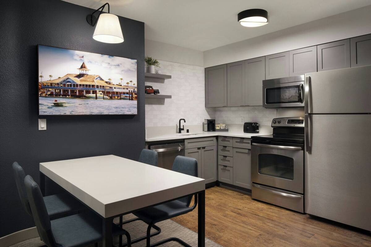 A modern kitchen with stainless steel appliances, a dining table with chairs, and a wall-mounted TV displaying a scenic image.
