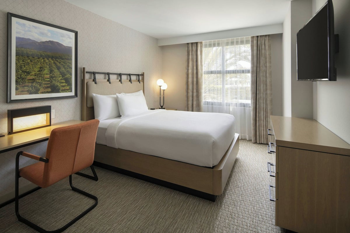 A hotel room with a bed, a desk with a chair, a TV, and a window with curtains.