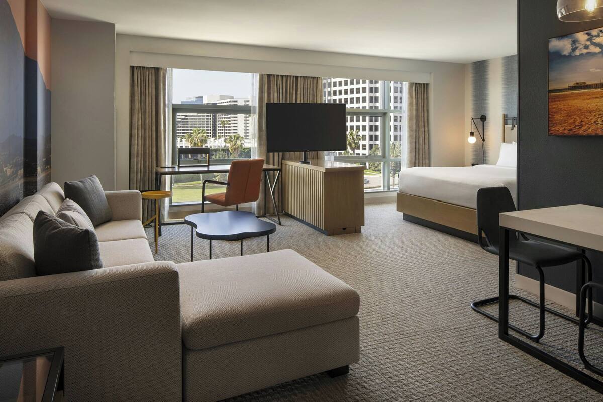 A hotel room with a sofa, a small table, a TV, a bed, and a view of the cityscape.