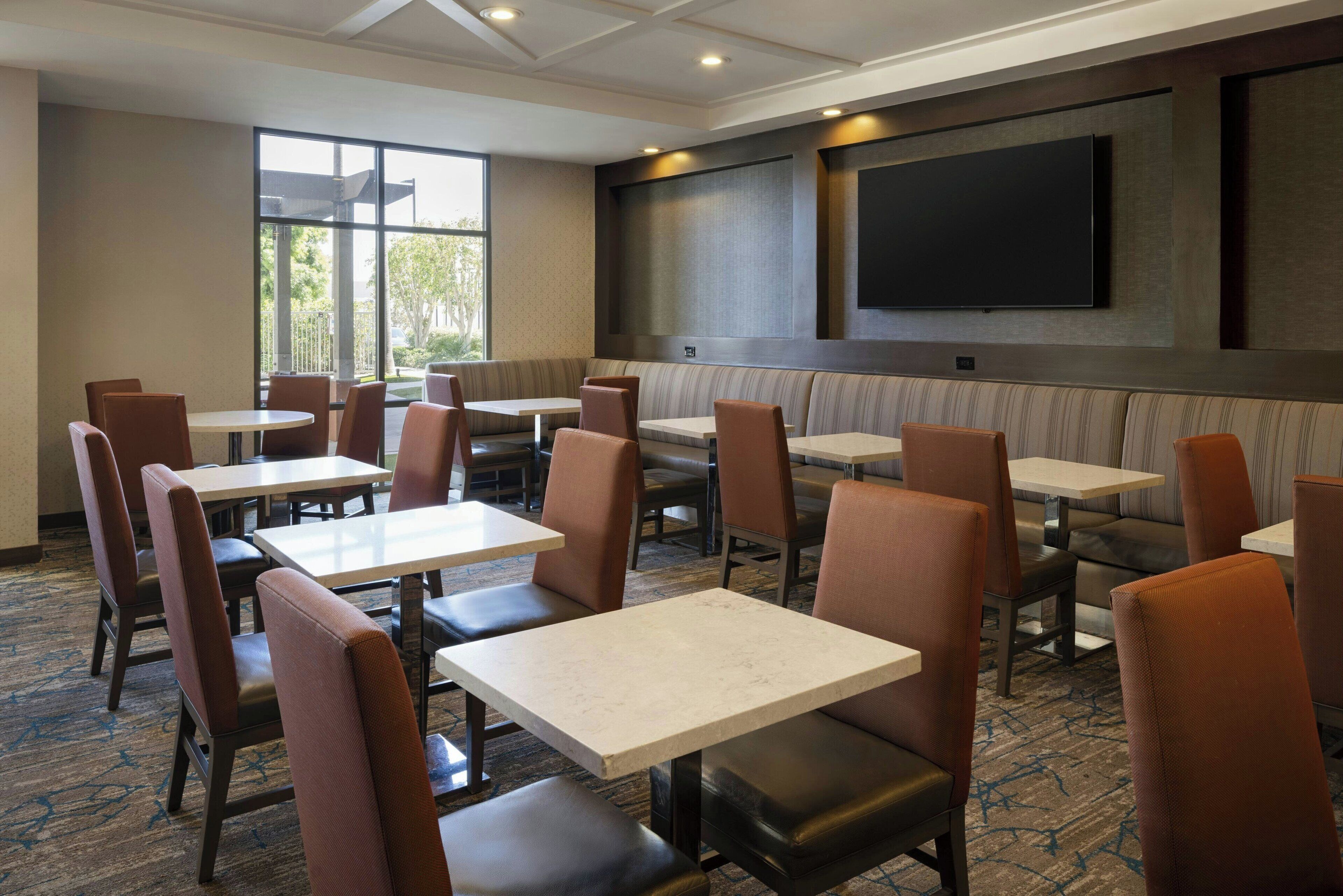 Een restaurantruimte in een hotel met zitjes en tafels, een flatscreen televisie aan de muur en grote ramen die zorgen voor natuurlijk licht.