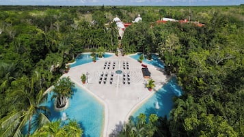 An aerial view of a resort with a central pool, surrounded by lush greenery and a beach area.