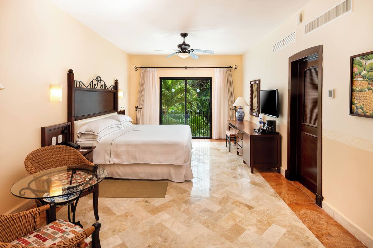 A hotel room with a bed, a glass table, a chair, a desk with a TV, and a door leading to a balcony with a view of greenery.