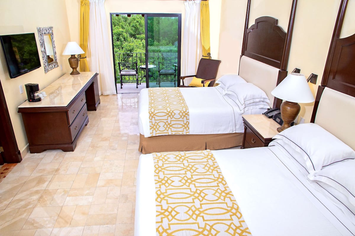 A hotel room with two beds, a TV, a desk with a lamp, and a balcony with chairs.