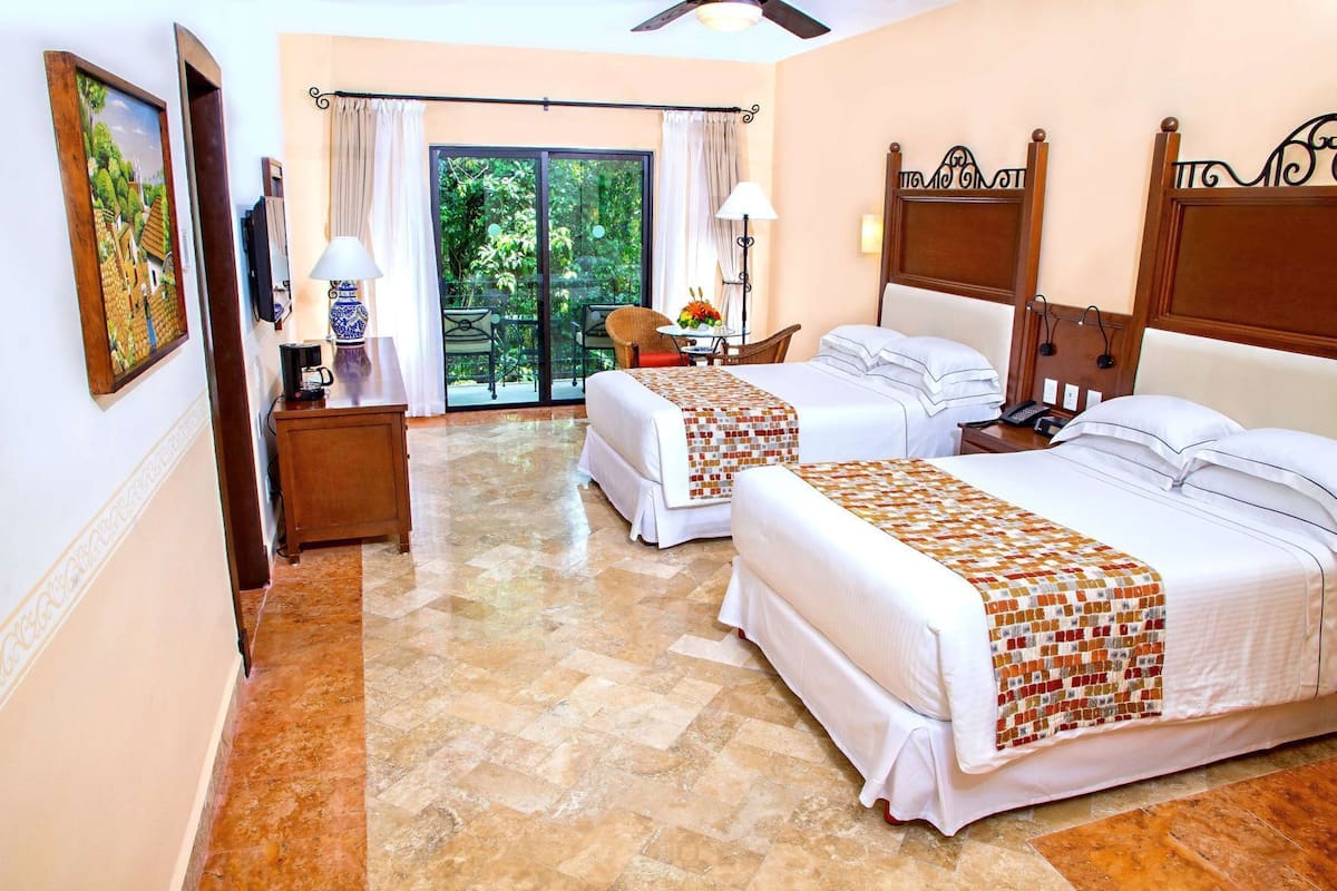 A hotel room with two beds, a coffee machine, a lamp, and a view of greenery through the glass door.